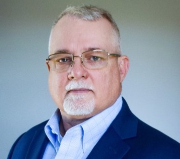 A middle-aged man in a suit jacket, white shirt and light blue tie. He appears to be posing for a photo, as he is facing t...